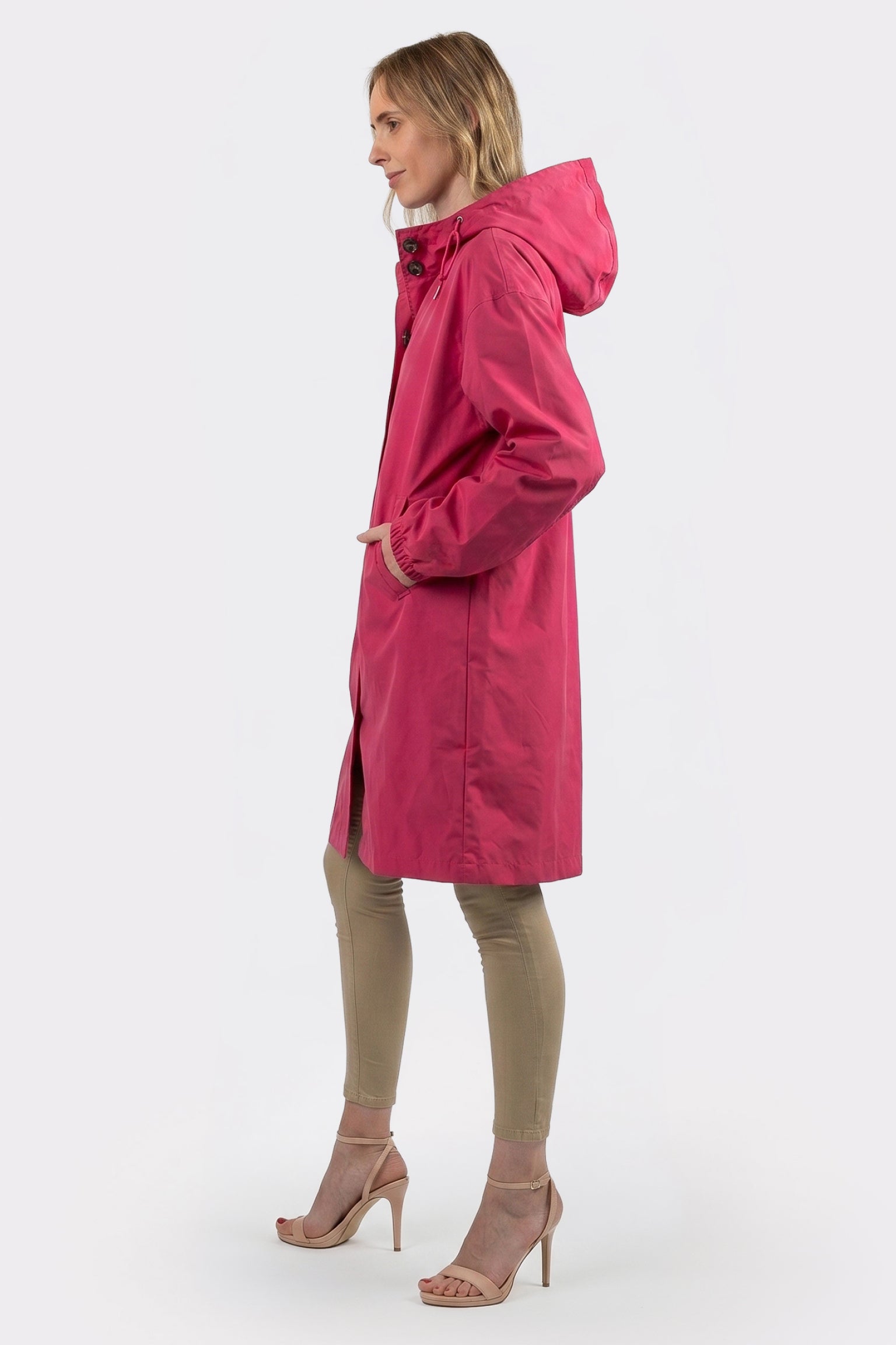 A person stands in profile wearing The Water Resistant Hooded Oversized Windbreaker Rain Jacket in bright pink, paired with beige pants and beige heeled sandals against a plain white background.