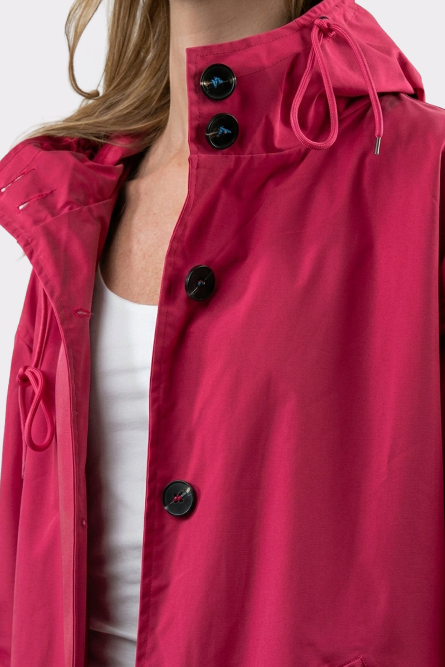 A person wearing the Icon Windbreaker Rain Jacket in bright pink with black buttons and a hood over a white top. The image displays the upper torso; part of the face is out of frame.