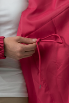 A person adjusts the drawstring of an Icon Windbreaker Rain Jacket in bright pink while wearing a white top.