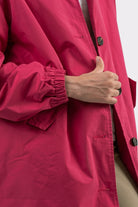 A person wearing the Icon Windbreaker Rain Jacket in bright pink with black buttons adjusts it with their right hand partially inside the front; the background is plain and light-colored.