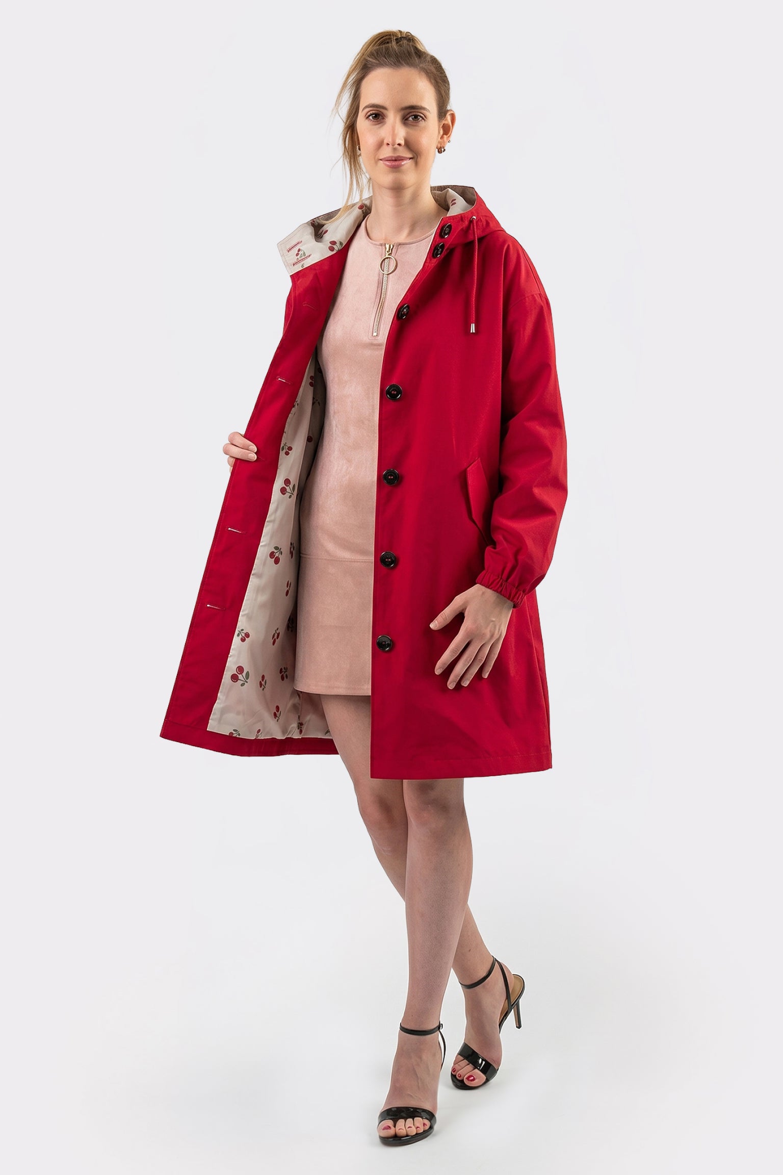 A woman faces forward against a plain white background, wearing The Water Resistant Hooded Oversized Windbreaker Rain Jacket in red, styled open to reveal its patterned lining over a light pink dress and black heeled sandals.