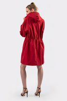 A person stands facing away, wearing the Icon Windbreaker Rain Jacket—a knee-length red hooded coat with an adjustable waist. They pair it with high-heeled, open-toe sandals against a plain light background.