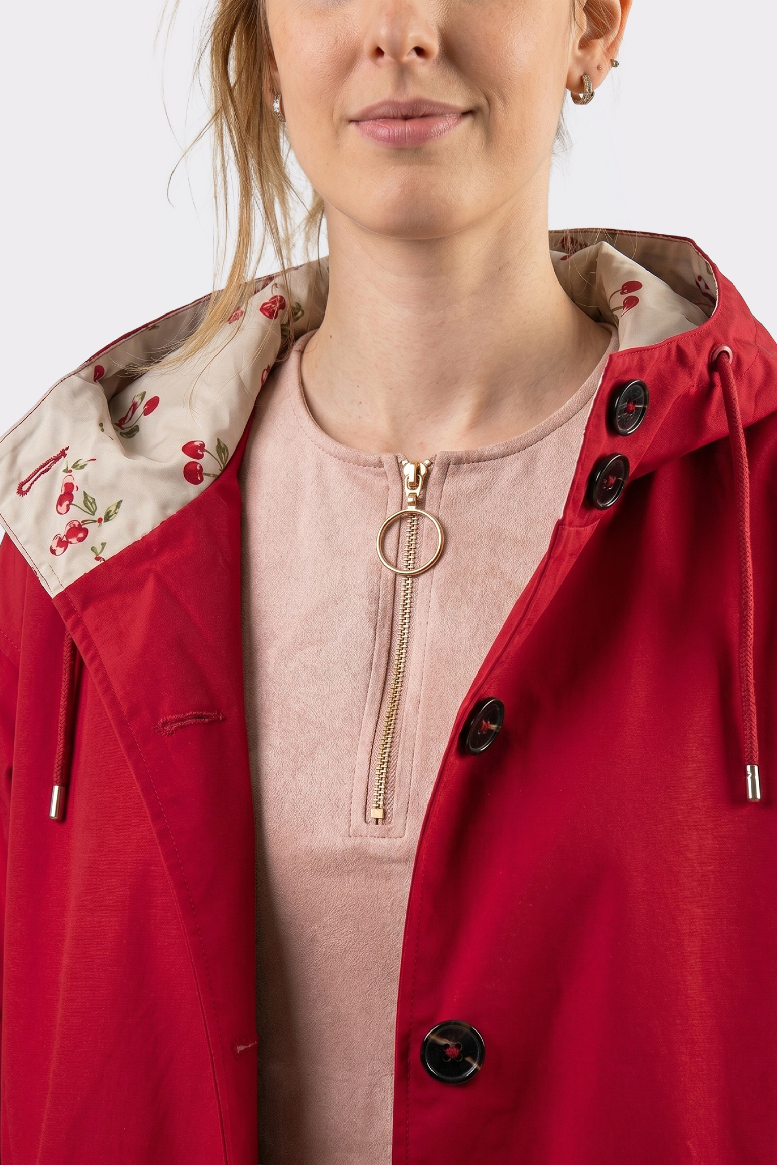 A woman wears The Water Resistant Hooded Oversized Windbreaker Rain Jacket in red with a cherry-patterned lining over a light pink top with a ring pull zipper; only her face and upper torso are shown.