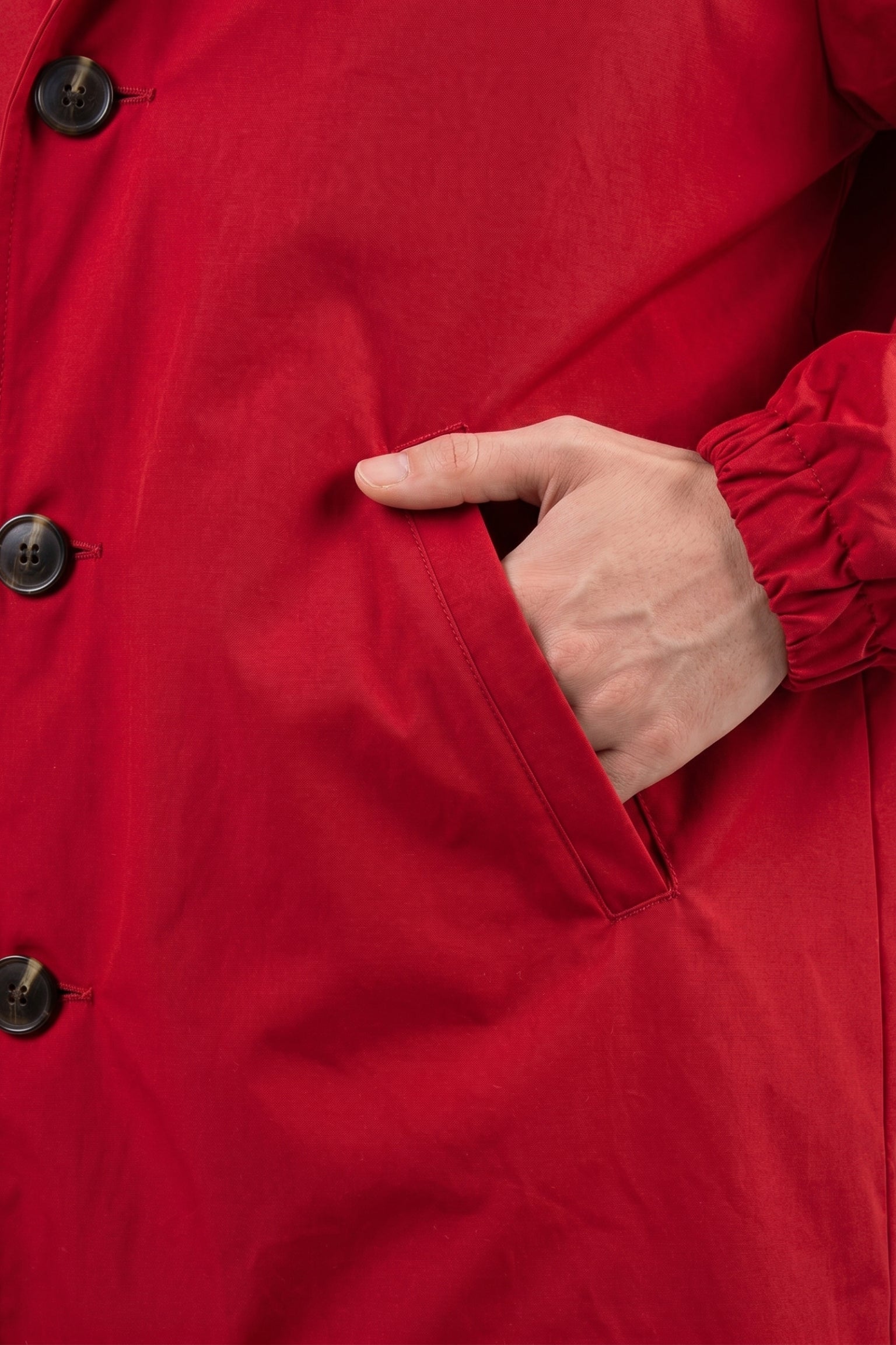 A close-up shows a person’s right hand in the pocket of The Water Resistant Hooded Oversized Windbreaker Rain Jacket, featuring its red color and black buttons.
