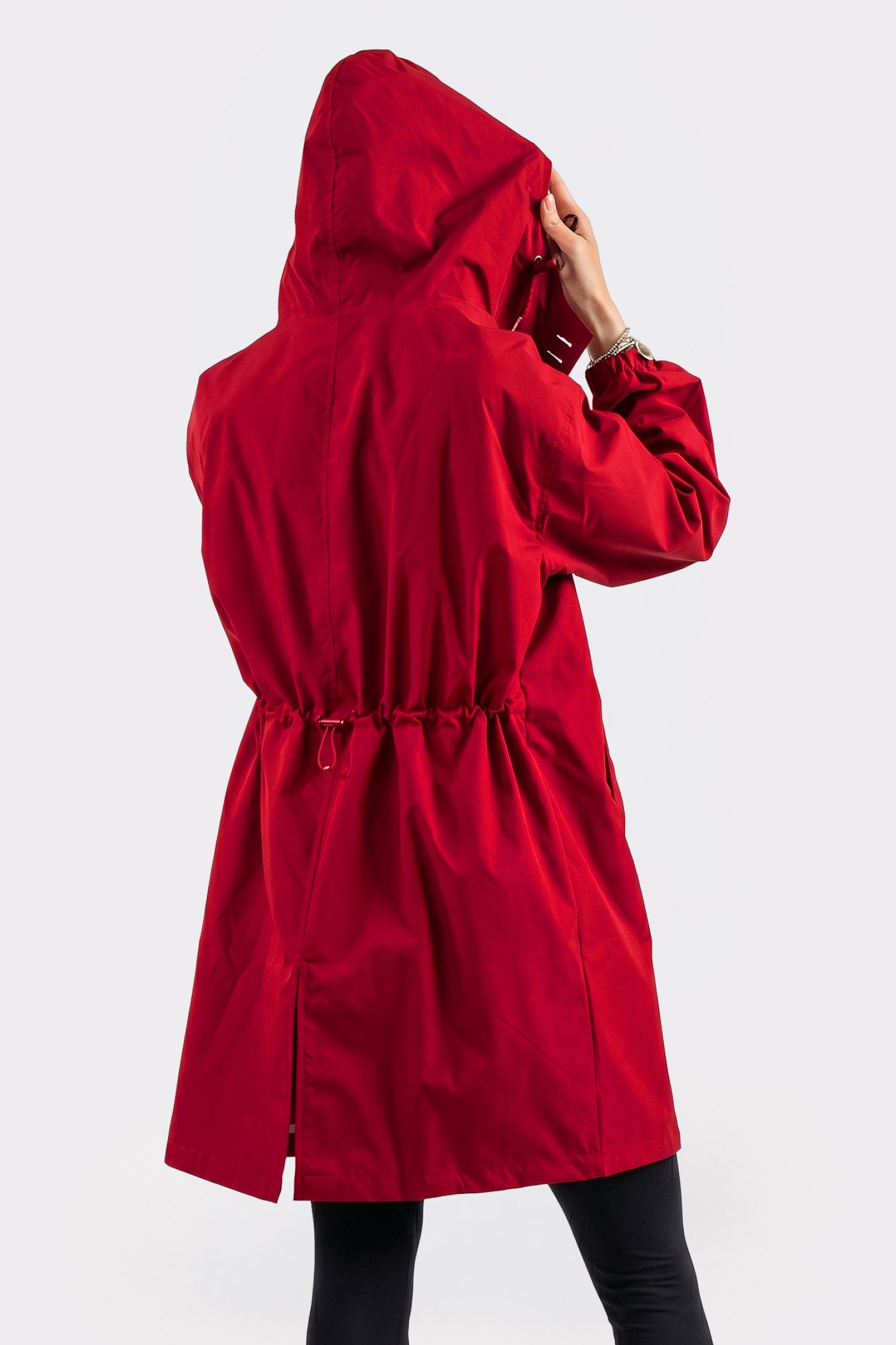 Wearing the Icon Windbreaker Rain Jacket, a person in a long, bright red hooded coat with a drawstring waist stands with their back to the camera, holding the hood. They also wear black pants.