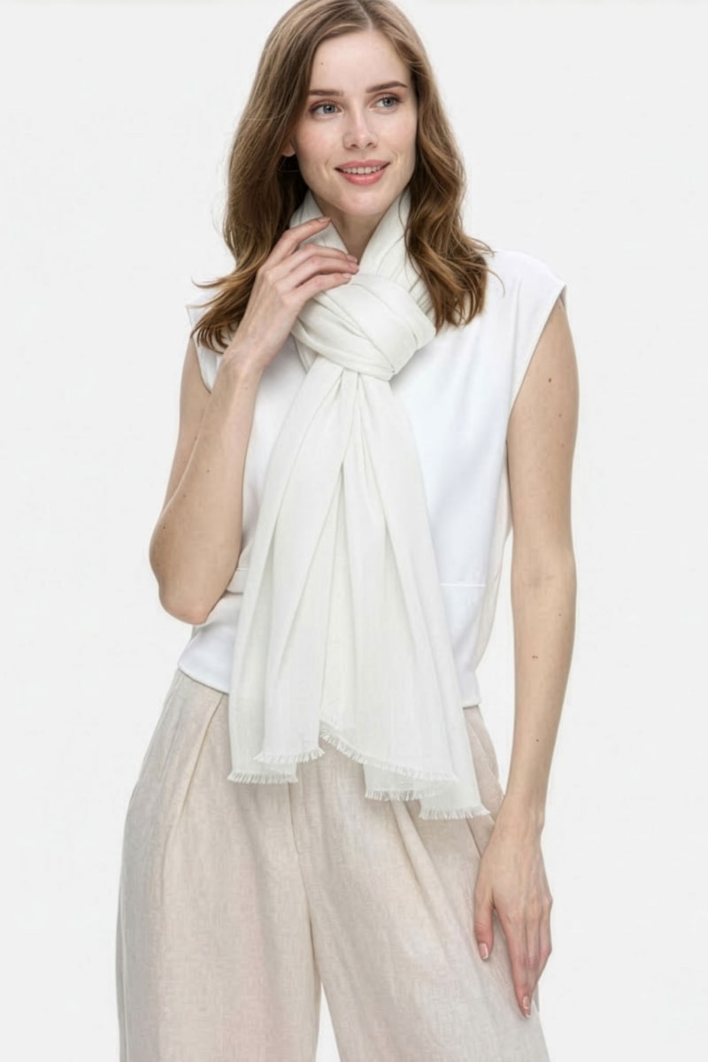 A woman with light brown hair wears a sleeveless white top, beige pants, and the looselyboho Ivory Whisper 100% Cashmere Scarf tied around her neck. She stands against a plain light background, looking slightly to the side and smiling softly.