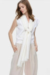 A woman stands against a plain background, hands in pockets, wearing a sleeveless white top, loose beige pants, and the looselyboho Ivory Whisper 100% Cashmere Scarf knotted at the front for a minimalist luxury look. She gazes downward.
