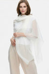A woman with light brown hair and a slight smile stands against a plain background, elegantly draped in the looselyboho Ivory Whisper 100% Cashmere Scarf over beige pants and a white top, capturing minimalist luxury.