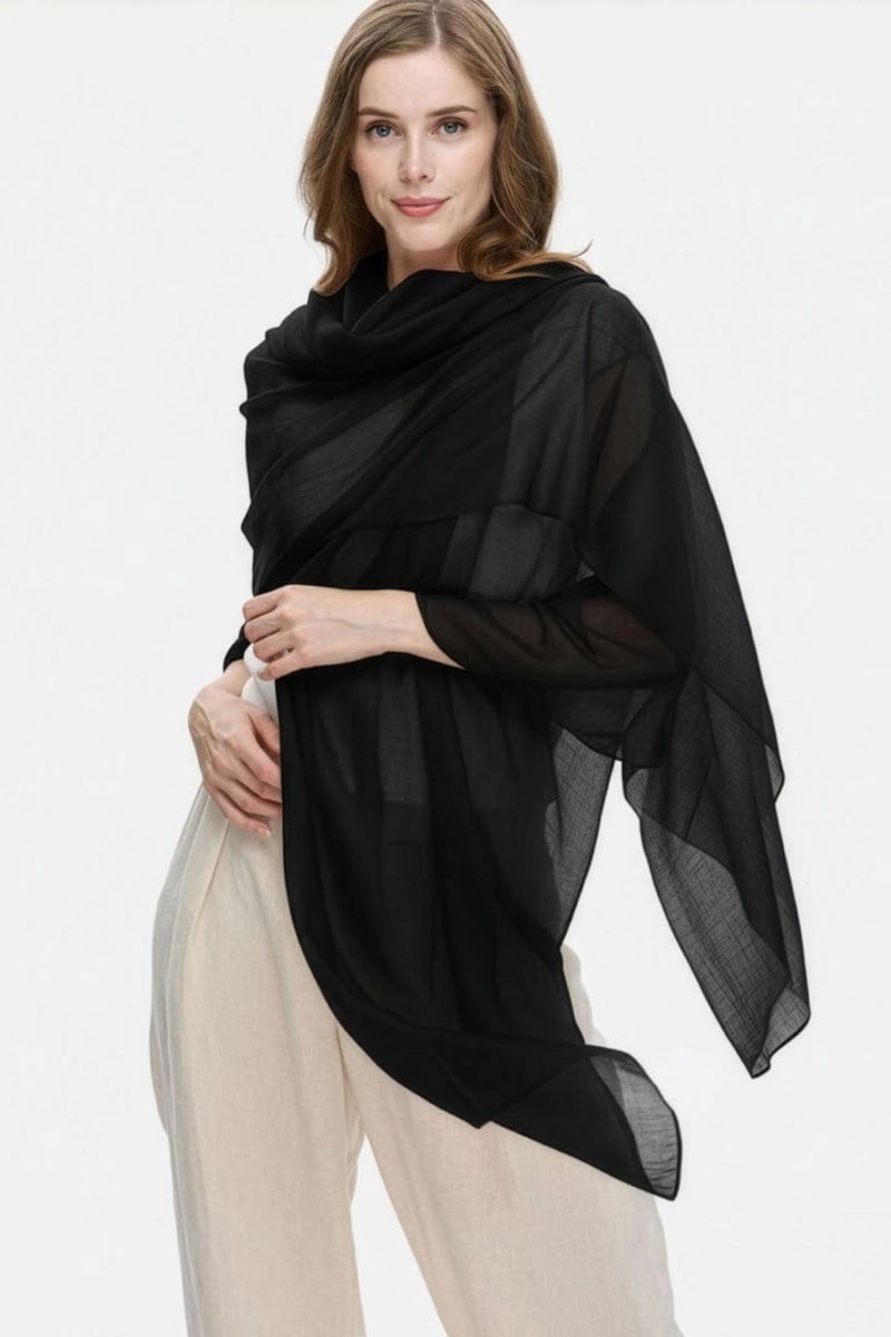 A woman with light brown hair wears the looselyboho Noir Eclipse 100% Cashmere Scarf draped over her shoulders and arms, paired with loose, light-colored pants, standing against a plain white background.