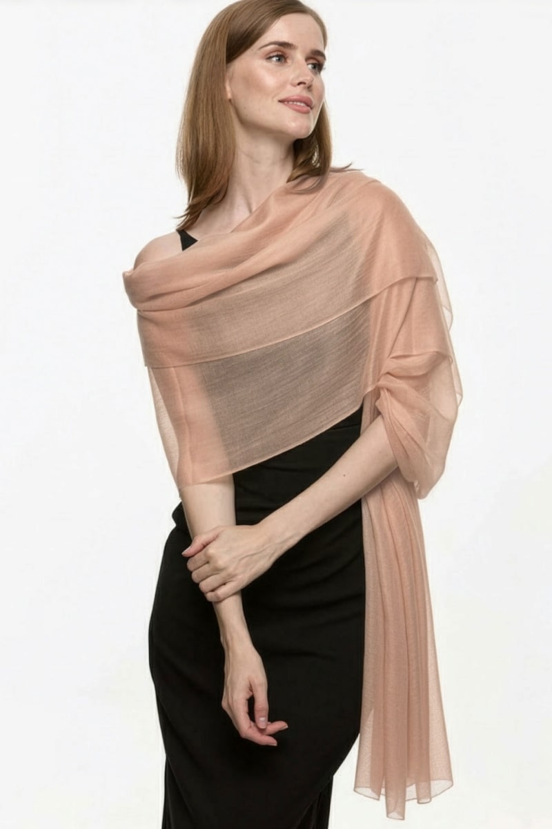 A woman with light brown hair wears the looselyboho Sandstone Dune 100% Cashmere Scarf, styled over her shoulders with a sleeveless black dress, standing against a plain white background and gazing slightly to the side.