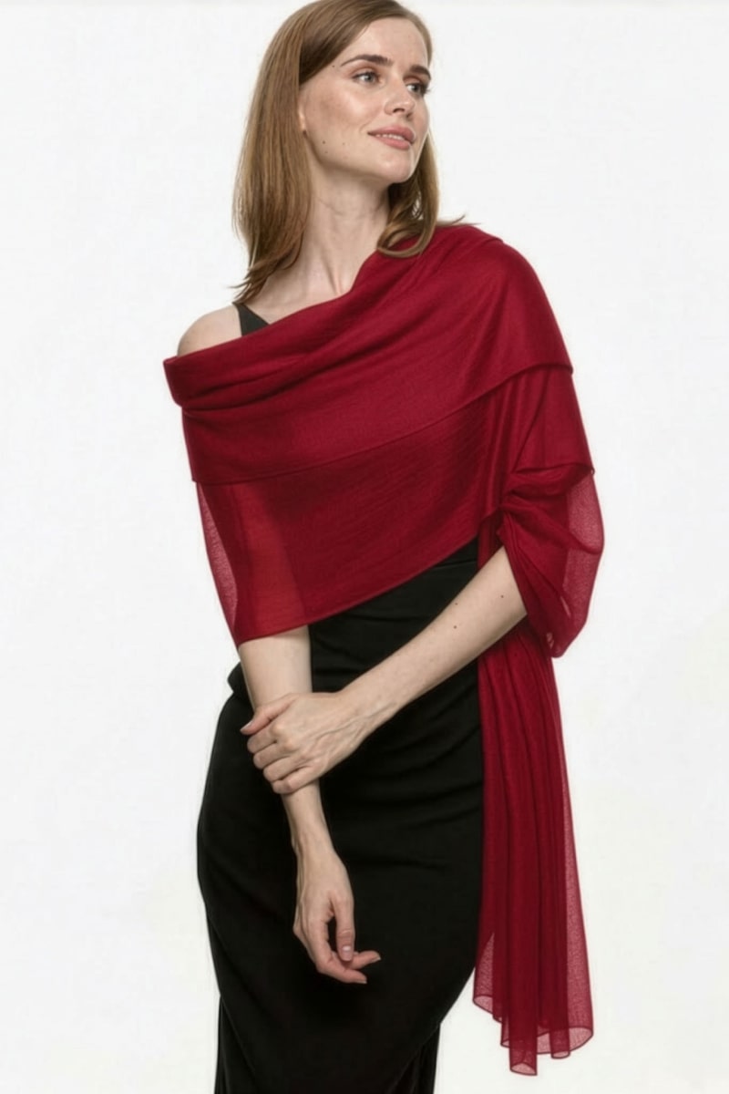 A woman with light brown hair stands against a white background, wearing a black dress and the looselyboho Velvet Merlot 100% Cashmere Scarf draped over her shoulders, looking slightly to the side with a neutral expression.