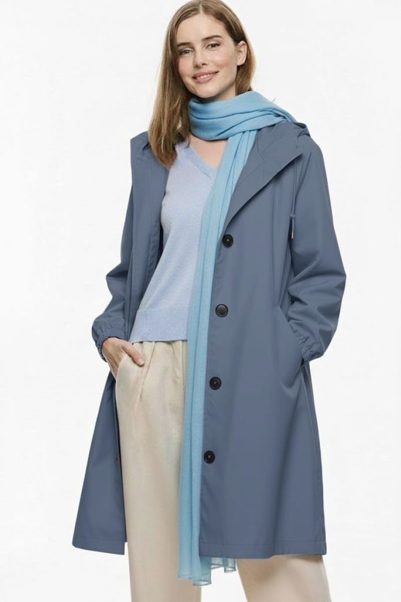 A woman poses against a plain background, smiling slightly with one hand in her pocket. She wears a blue coat, a light blue top, beige pants, and the looselyboho Sky Mist 100% Cashmere Scarf draped lightly around her neck.