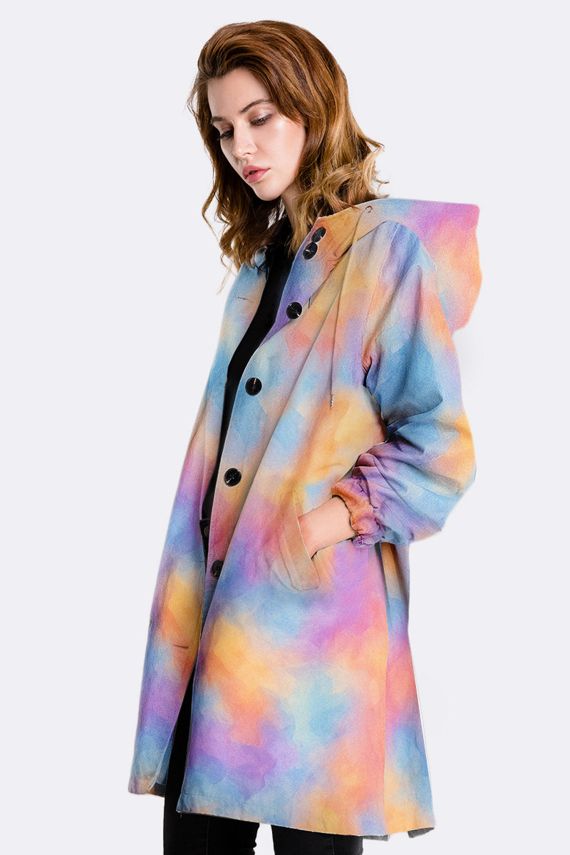 A woman with light brown hair wears the looselyboho CANVAS SKY WINDBREAKER, a pastel rainbow tie-dye hooded coat with black buttons. She looks down with a neutral expression against a plain light background.