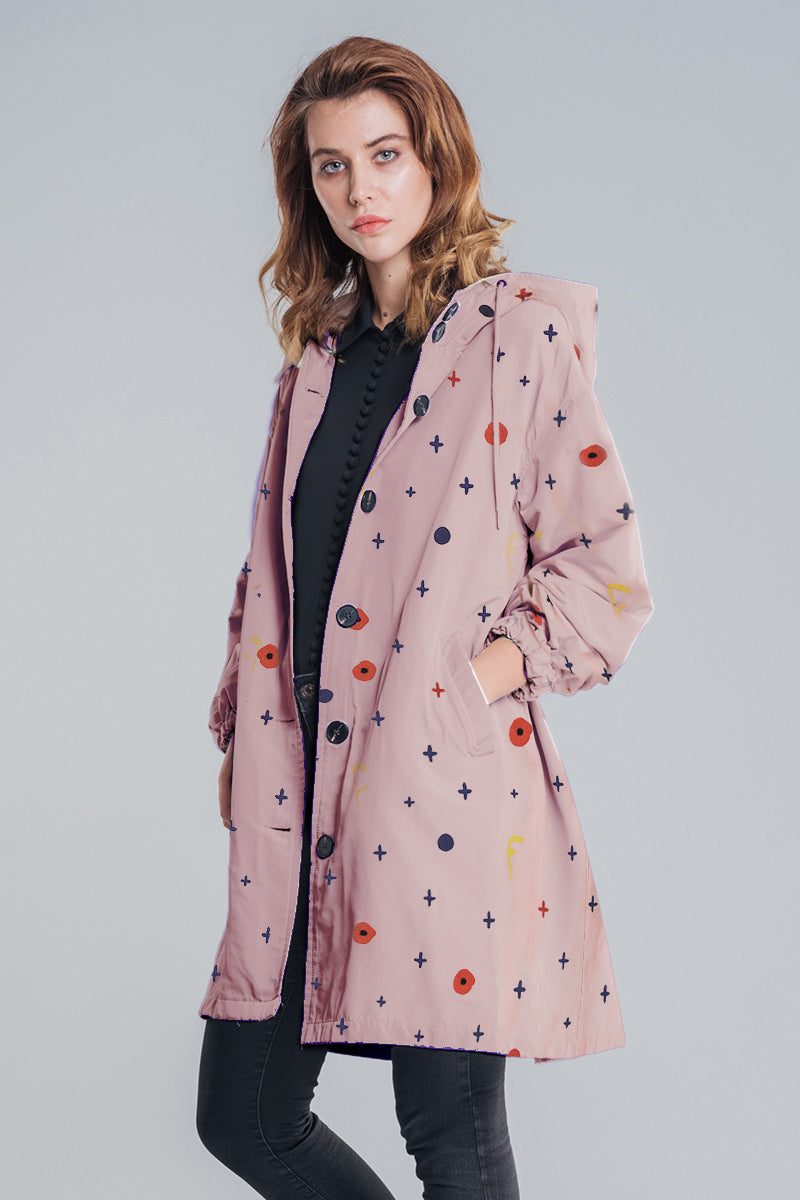 A woman with light brown hair and a neutral look faces forward wearing the DOT & BLOOM WINDBREAKER by looselyboho—a light pink, water-resistant jacket with black buttons and a geometric pattern over a black outfit.