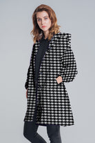 A person stands against a plain background wearing the looselyboho HOUNDSTOOTH WINDBREAKER, featuring a black and white pattern, pockets, and a hood, paired with a black top and dark pants.