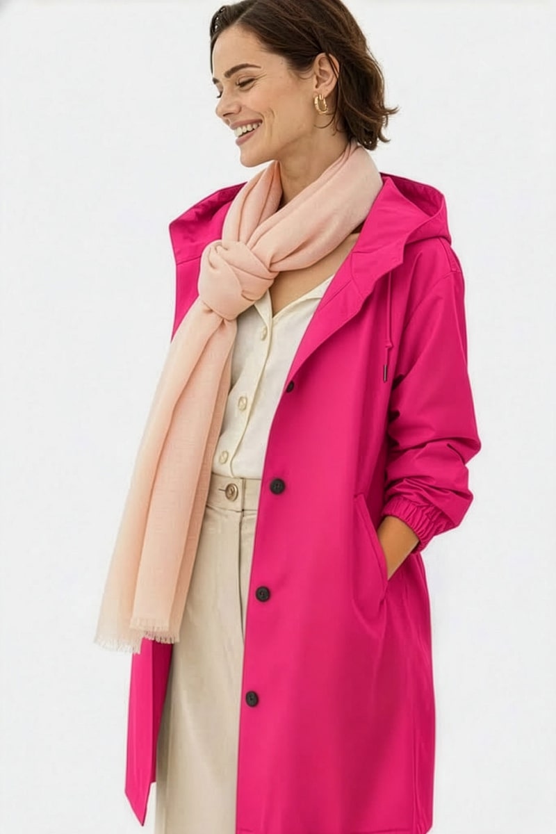 A person smiles with hands in pockets, wearing a bright pink coat and the looselyboho Blush Rose 100% Cashmere Scarf, styled over a light outfit, against a plain white background.